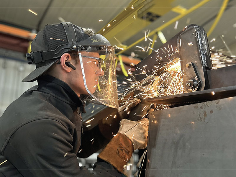 Fabricator in a face shield grinding a steel dump body component in the Rugged shop