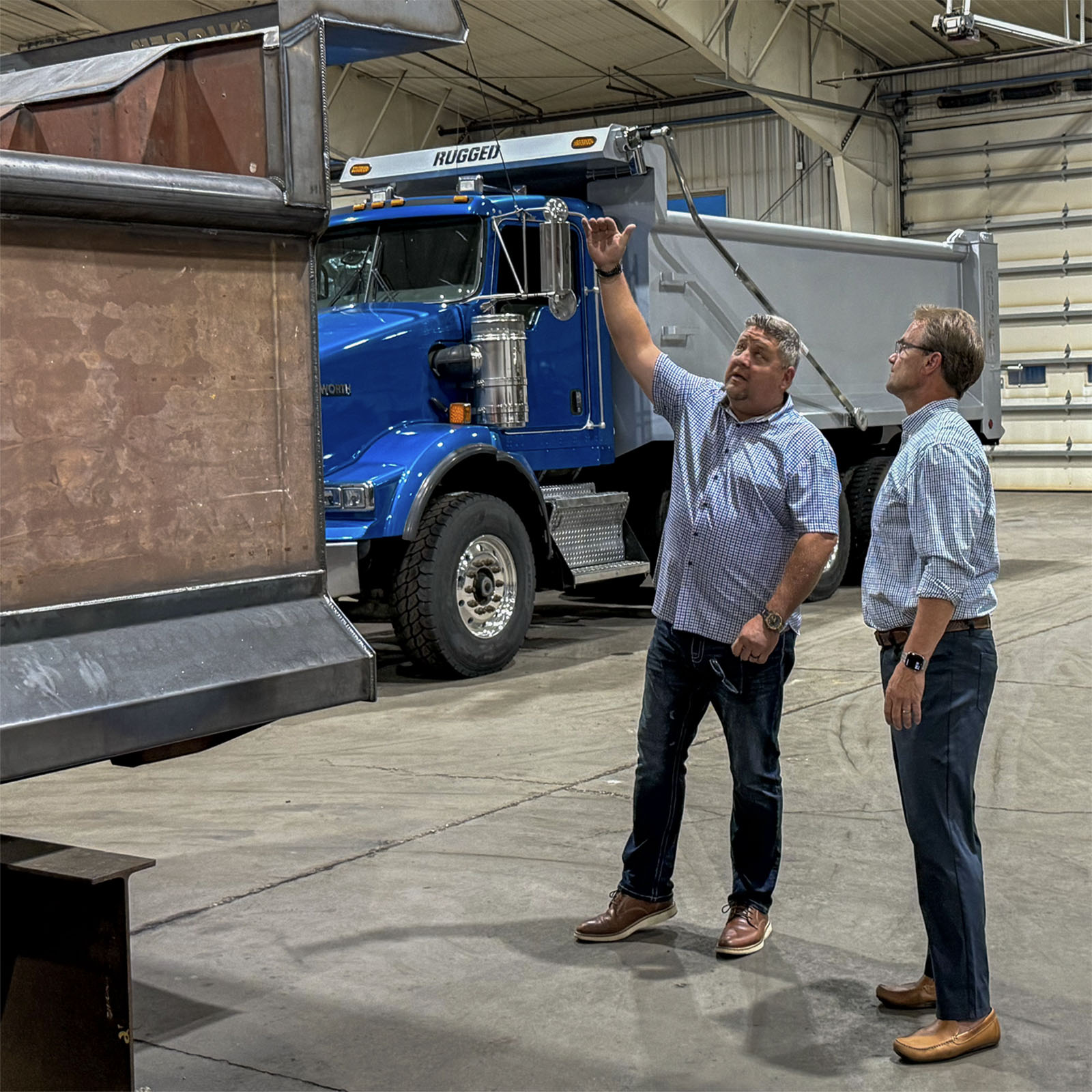 Two men reviewing a raised dump body in the Rugged shop, with a blue Rugged dump truck in the background