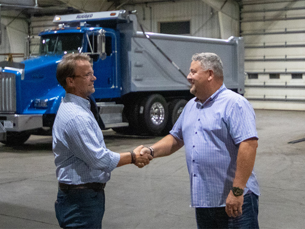 Two men shaking hands in front of a blue truck with a finished Rugged dump body in the shop
